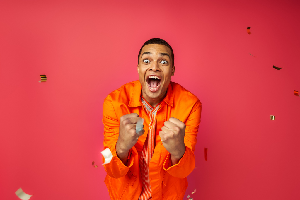 nadšený africký Američan v oranžové košili křičí a ukazuje vítězství gesto blízko konfety na červené - Fotografie, Obrázek