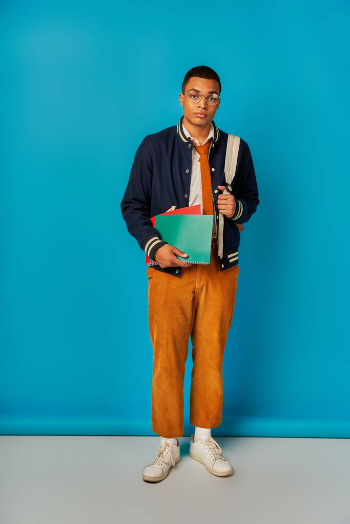 stylish african american student with backpack and notebooks looking at camera on blue background - Photo, Image