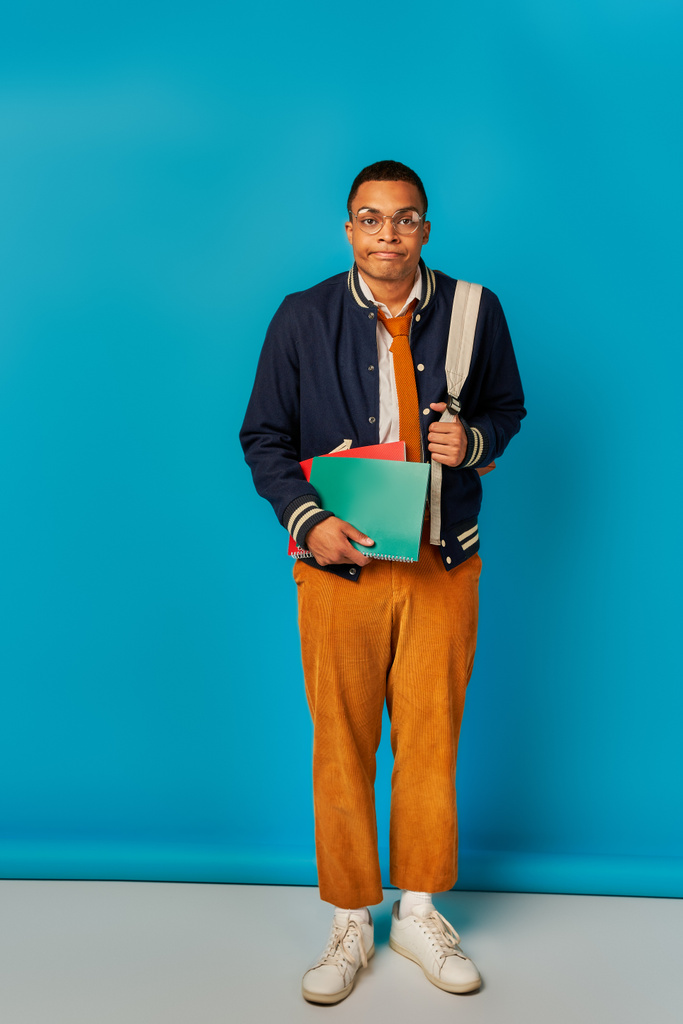 bored african american student with notebooks and backpack puffing cheeks on blue background - Photo, Image