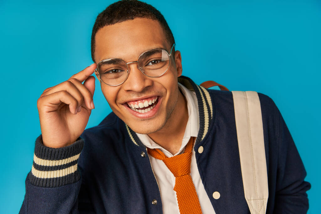 joyeuse étudiante afro-américaine en veste ajustant les lunettes, regardant la caméra sur bleu, portrait - Photo, image