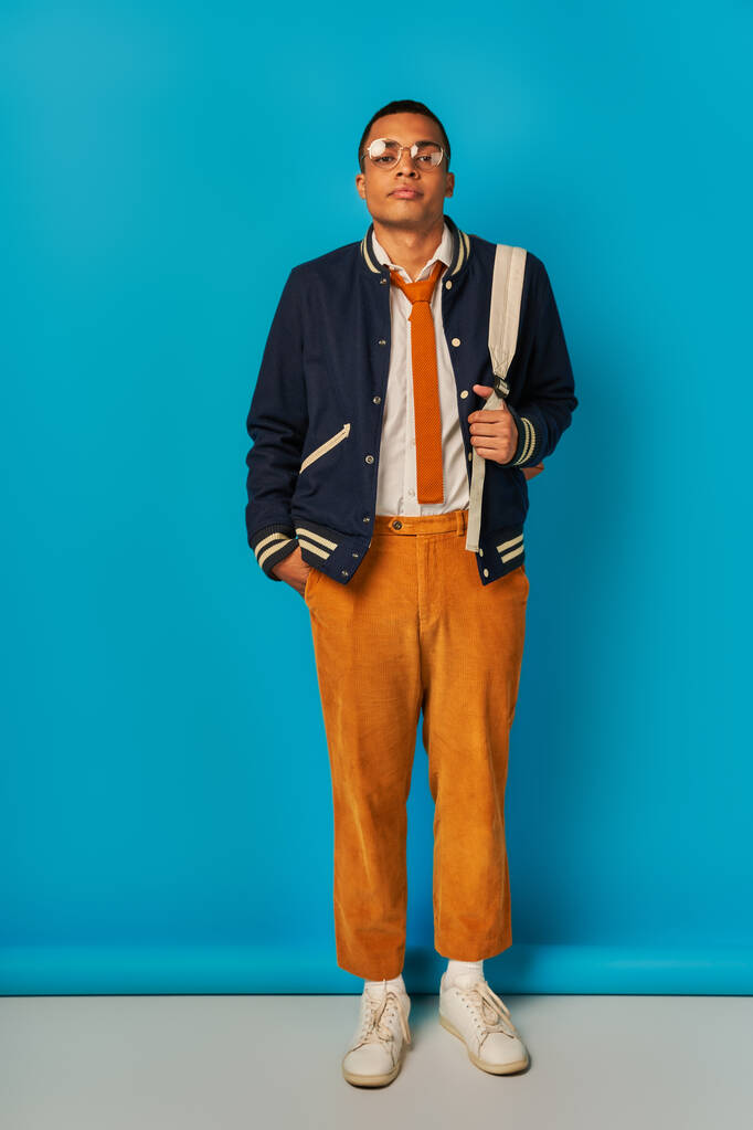 estudiante afroamericano confiado en chaqueta y pantalones anaranjados tomados de la mano en el bolsillo en azul - Foto, Imagen