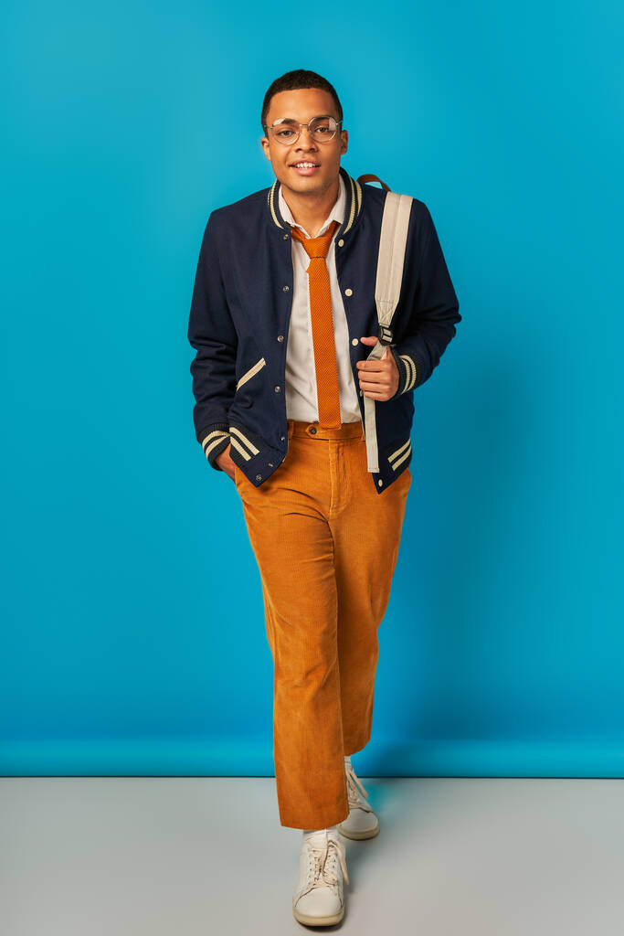 стильний афроамериканський студент в куртці і помаранчевих штанах, що йдуть рукою в кишені на синьому - Фото, зображення