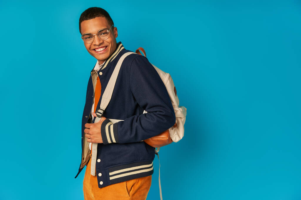 estudante afro-americano despreocupado em casaco e calça laranja, com mochila olhando para câmera em azul - Foto, Imagem