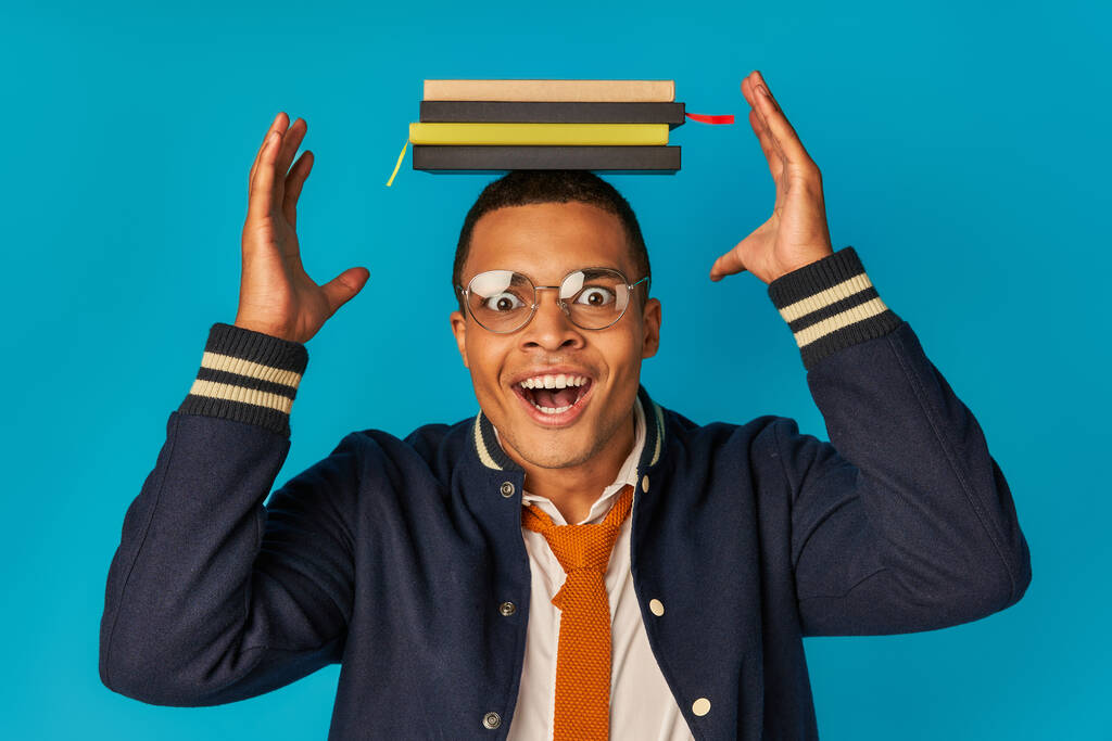 étudiant afro-américain étonné posant avec des cahiers sur la tête sur bleu, lunettes, veste élégante - Photo, image