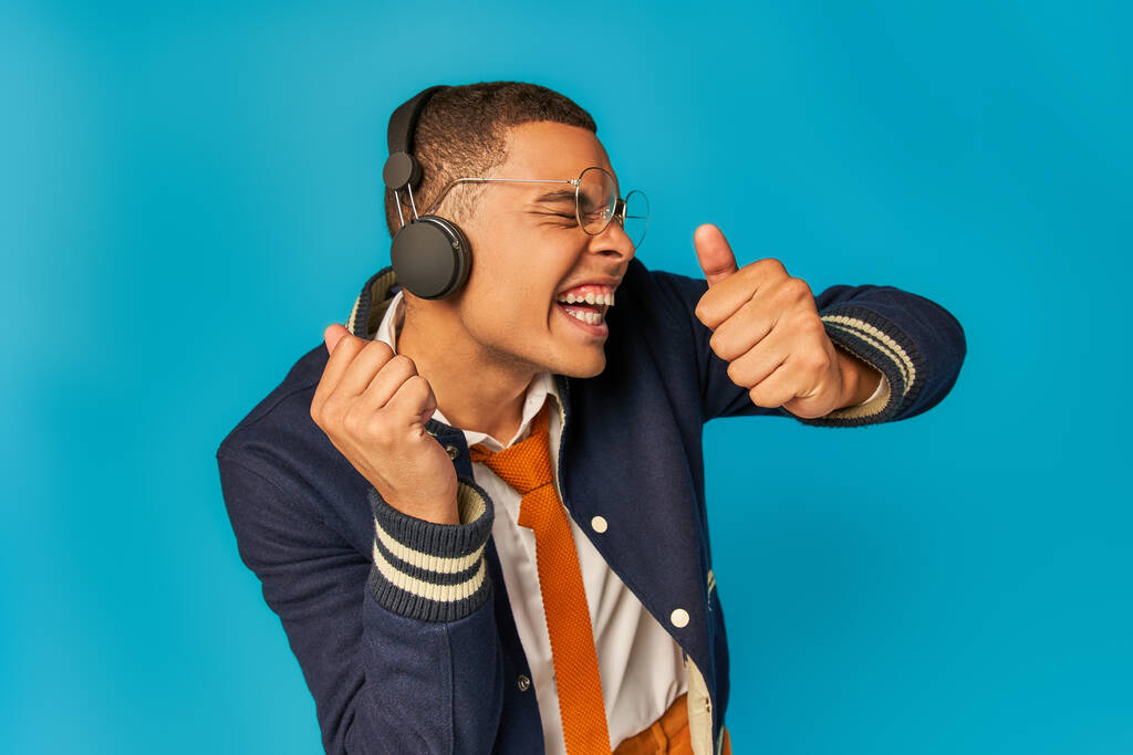 expresivo afroamericano estudiante escuchando música en auriculares y mostrando pulgares hacia arriba en azul - Foto, imagen