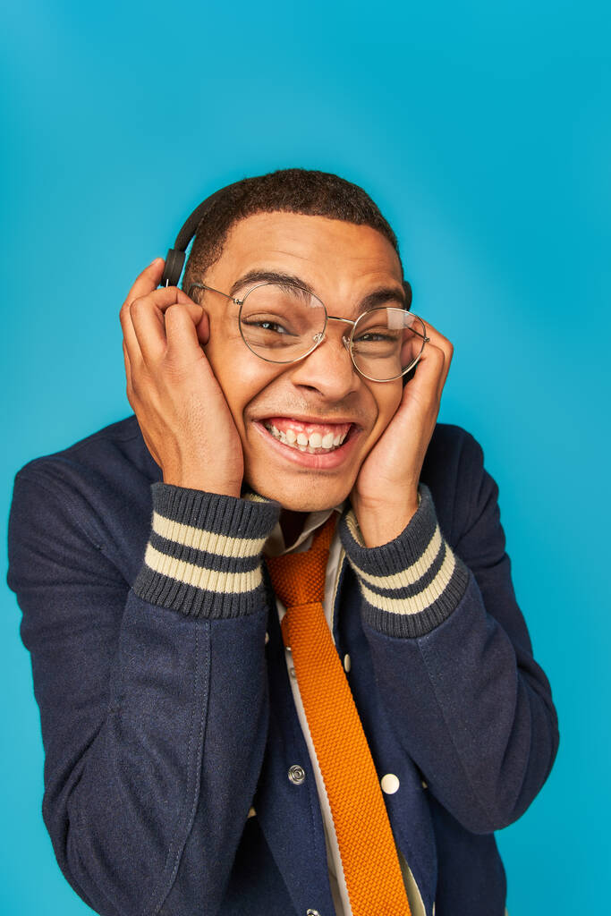radostný africký americký student v brýlích, bundě a sluchátkách při pohledu na kameru na modrou - Fotografie, Obrázek