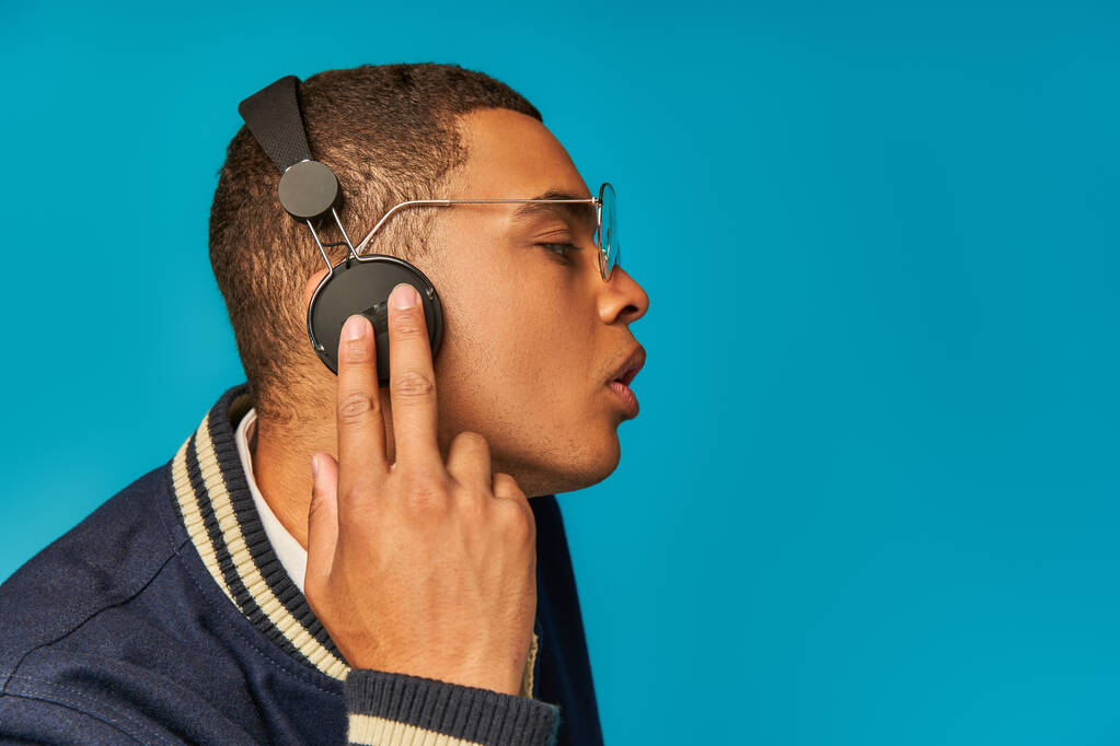 προφίλ του Αφροαμερικανού φοιτητή με σακάκι και γυαλιά ηλίου που αγγίζει ασύρματα ακουστικά στο μπλε - Φωτογραφία, εικόνα