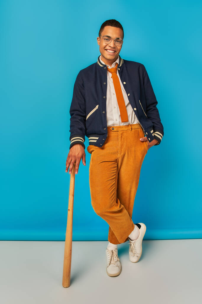 glimlachende Afro-Amerikaanse student in jas en oranje broek poseren met honkbalknuppel op blauw - Foto, afbeelding