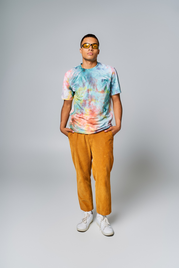 Afrykański Amerykanin w okularach przeciwsłonecznych, koszulce z krawatem i pomarańczowych spodniach z rękami w kieszeniach na szarości - Zdjęcie, obraz