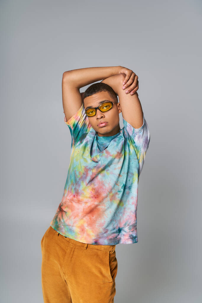 stylish african american guy in sunglasses and tie-dye t-shirt with hands above head on grey - Photo, Image