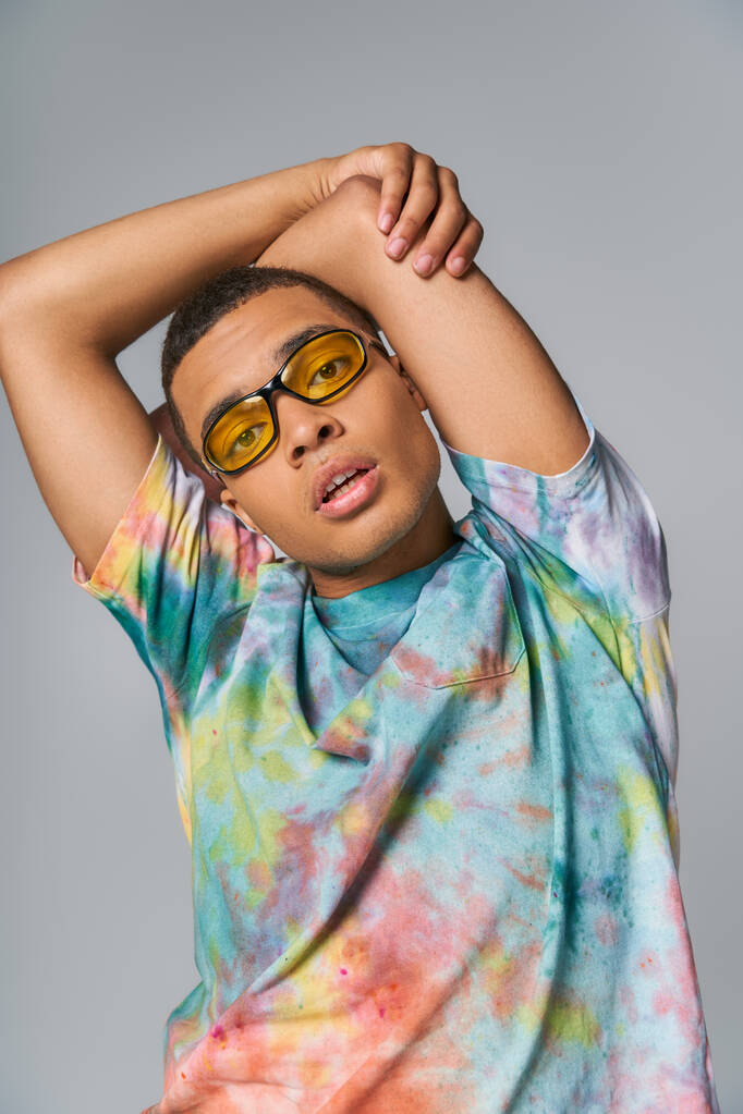 african american man with hands above head looking at camera on grey, sunglasses, tie-dye t-shirt - Photo, Image
