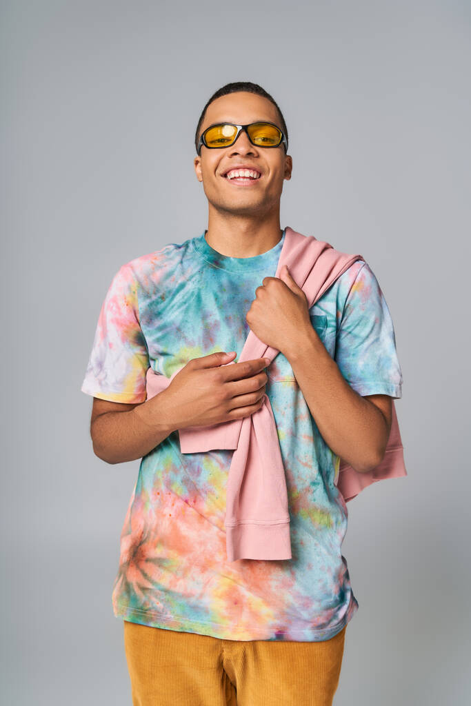 happy african american man in sunglasses and tie-dye t-shirt looking at camera on grey - Photo, Image