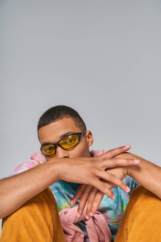 jong Afrikaans amerikaans man in trendy zonnebril en tie-dye t-shirt kijken naar camera op grijs - Foto, afbeelding