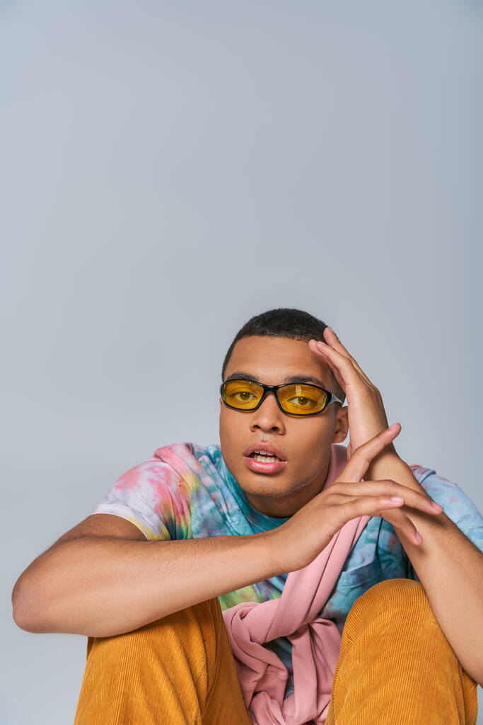 молодой африканский американец в модных солнцезащитных очках и футболке с красителем, смотрящий в камеру на сером - Фото, изображение