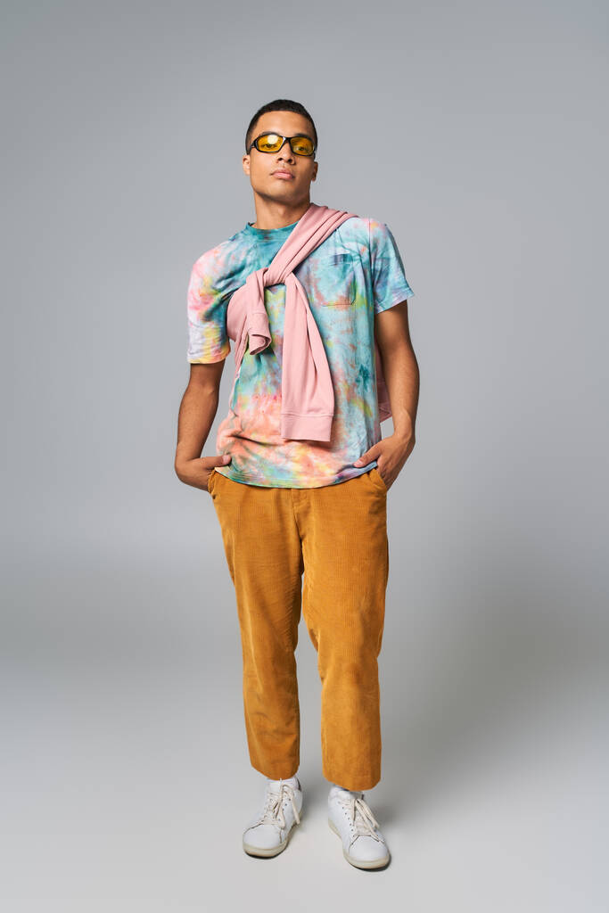 confident african american man, orange pants, sunglasses, tie-dye t-shirt, hands in pocket, on grey - Photo, Image