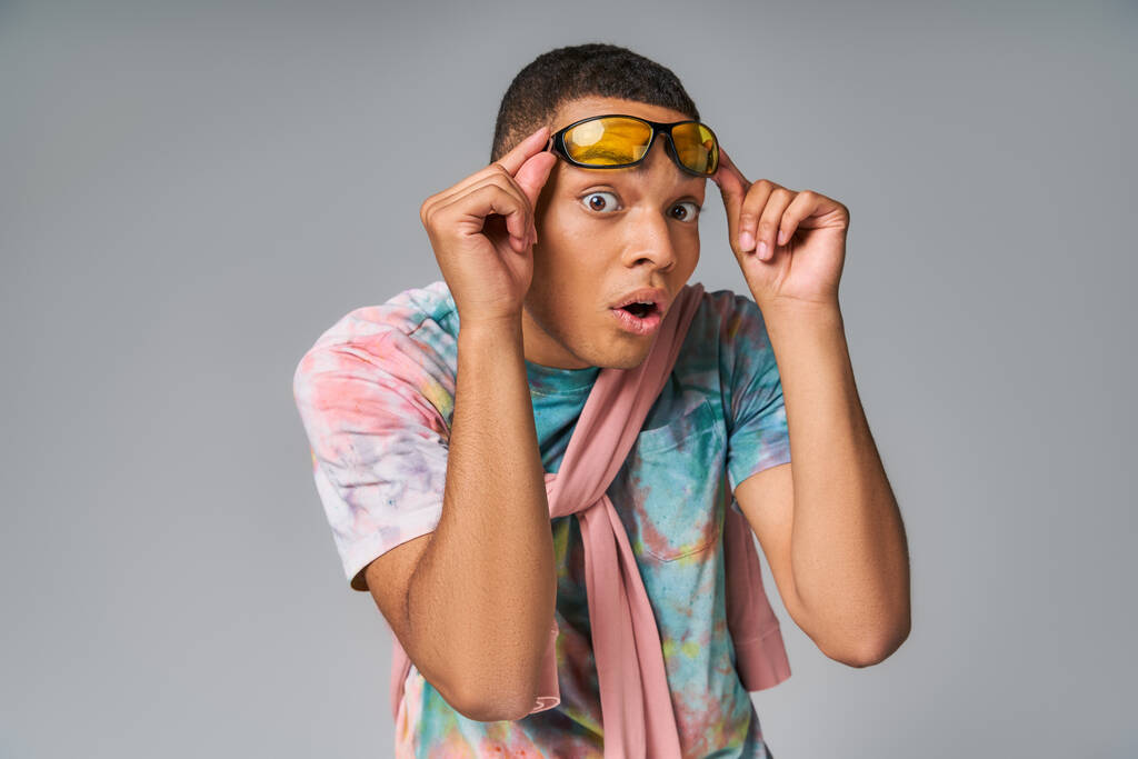 surpreso afro-americano cara em tie-dye camisa segurando óculos de sol e olhando para a câmera em cinza - Foto, Imagem