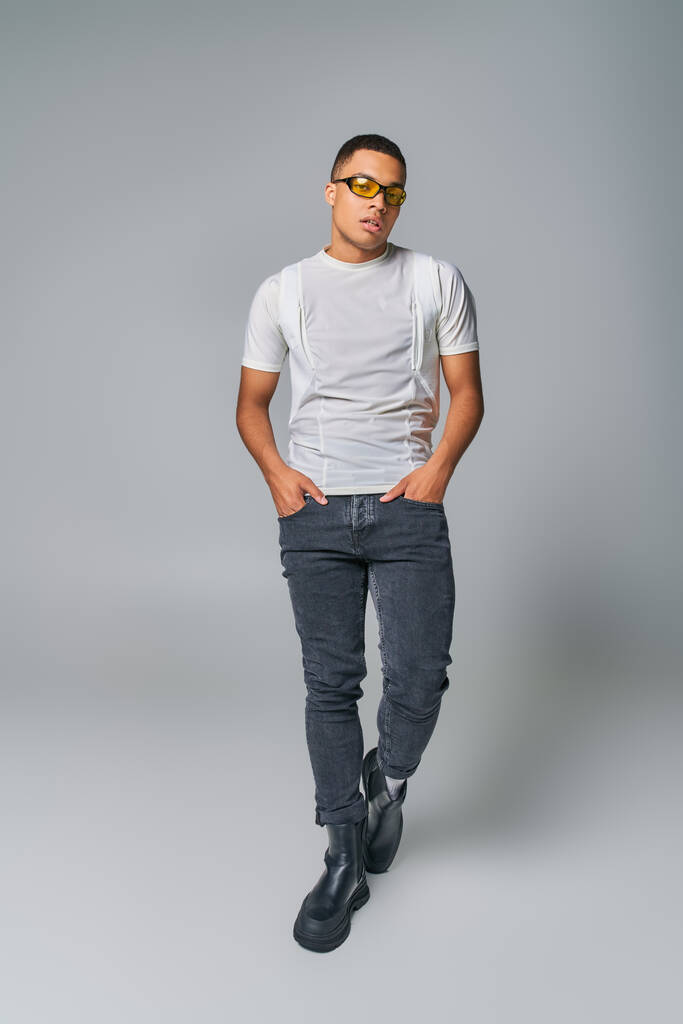 stylowy Afrykański Amerykanin w okularach przeciwsłonecznych, t-shirt, dżinsy patrzące w kamerę na szarości - Zdjęcie, obraz