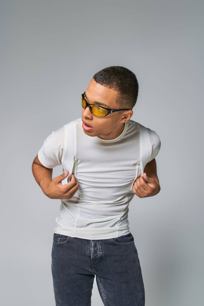 jeugdige Afro-Amerikaanse man in stijlvolle t-shirt en zonnebril weg te kijken op grijs - Foto, afbeelding