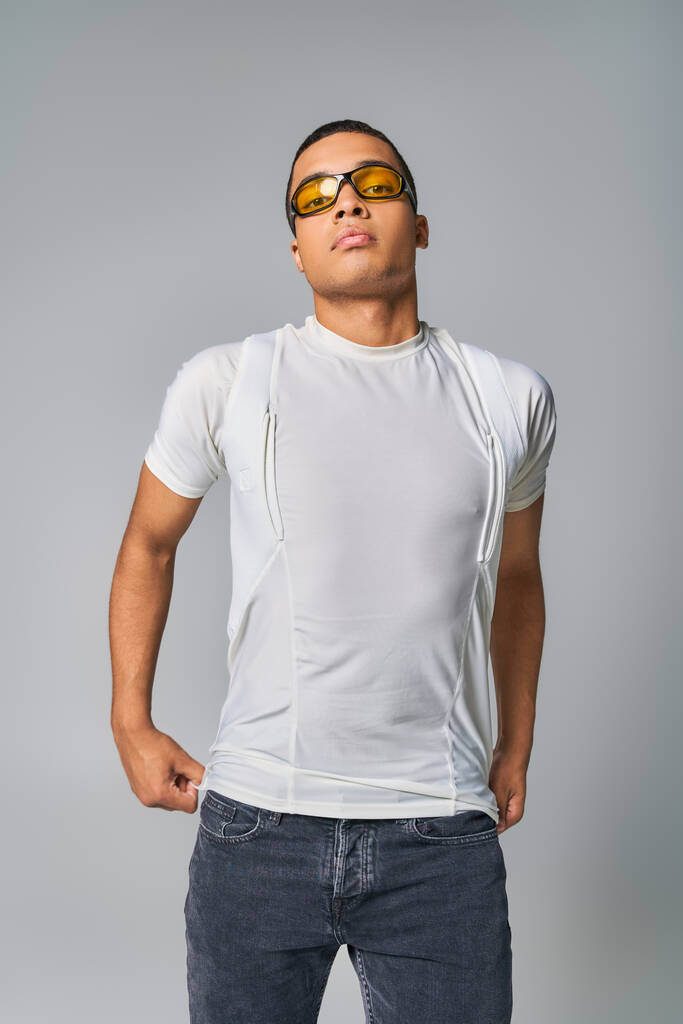 moda moderna, cara americano africano em t-shirt, jeans e óculos de sol olhando para a câmera em cinza - Foto, Imagem