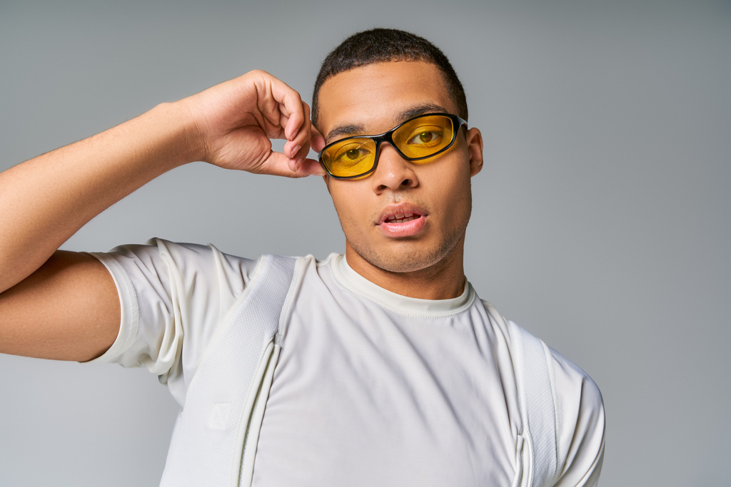 young, stylish african american man in t-shirt adjusting sunglasses, looking at camera on grey - Photo, Image