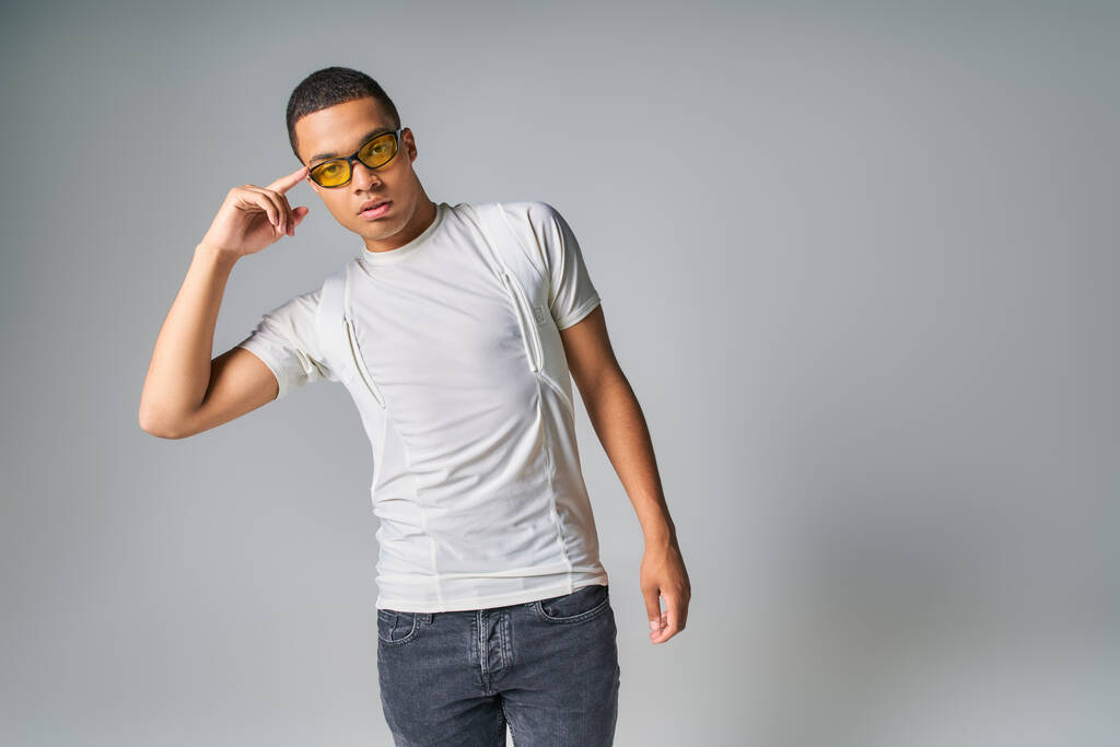 homme afro-américain à la mode en t-shirt, jeans et lunettes de soleil pointant la tête sur gris - Photo, image