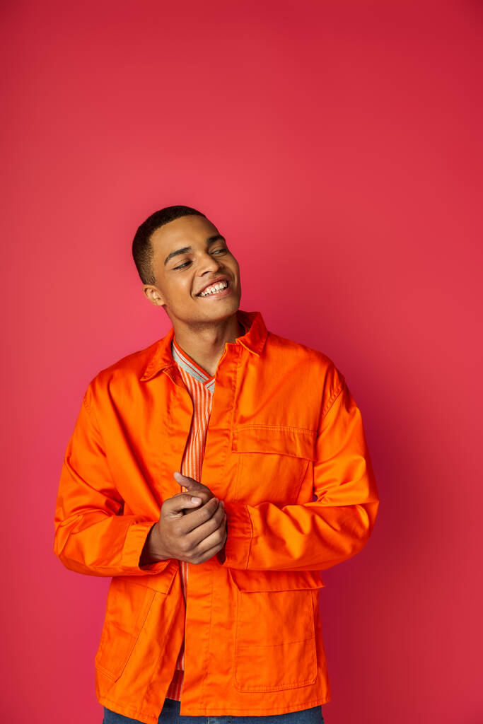 vrolijk en trendy Afrikaans amerikaanse man in oranje shirt weg te kijken op rode achtergrond - Foto, afbeelding