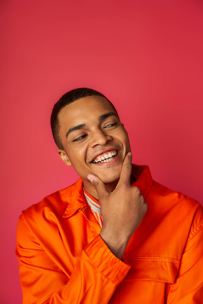 sonriente y elegante hombre afroamericano en camisa naranja tocando la cara y mirando hacia otro lado en rojo - Foto, Imagen