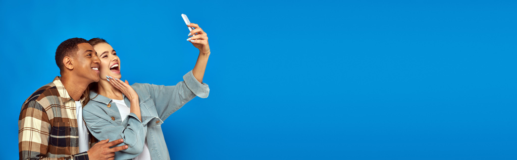 cheerful interracial couple taking selfie while looking at smartphone on blue backdrop, banner - Photo, Image