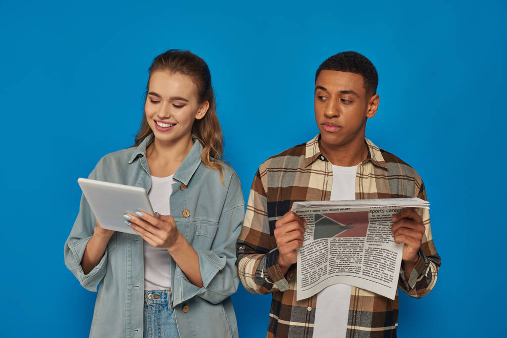 Afrikkalainen amerikkalainen mies lukee painettua sanomalehteä, kun blondi nainen käyttää tablettia sinisellä pohjalla - Valokuva, kuva