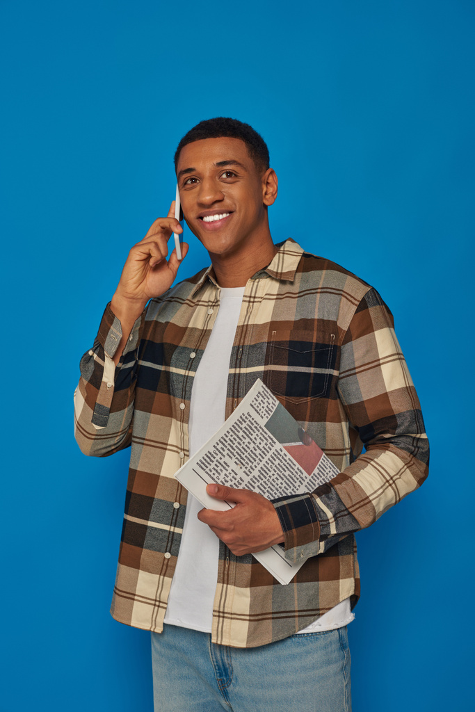glücklicher afrikanisch-amerikanischer Mann in kariertem Hemd, der telefoniert und Zeitung vor blauem Hintergrund hält - Foto, Bild