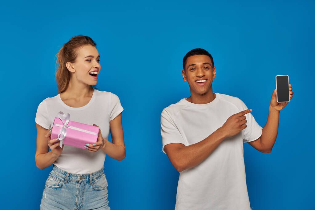 E-ticaret, mutlu Afro-Amerikalı adam mavi arka planda hediyeli bir kadının yanındaki akıllı telefonu işaret ediyor. - Fotoğraf, Görsel