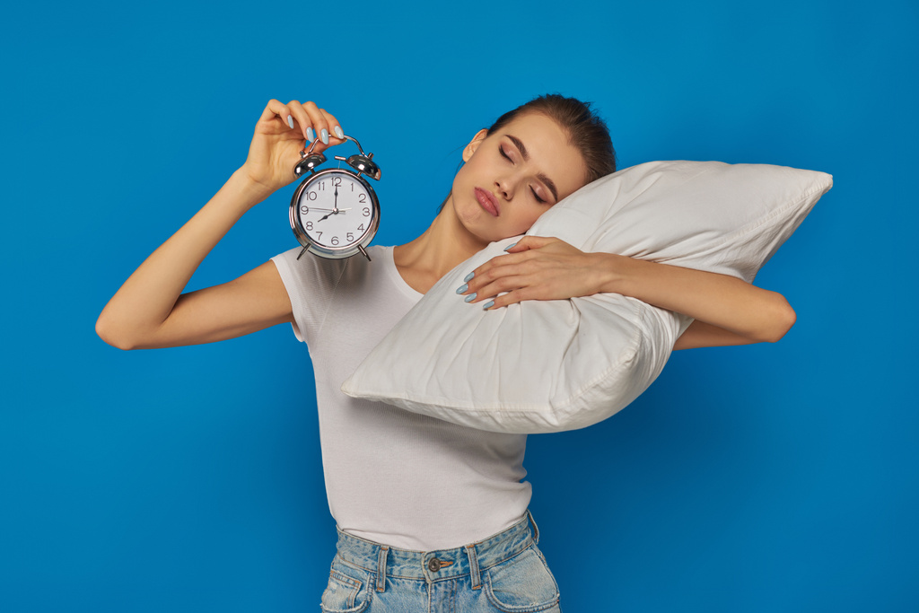 недовольная молодая женщина держит винтажный будильник и лежит на подушке на синем фоне, утро - Фото, изображение
