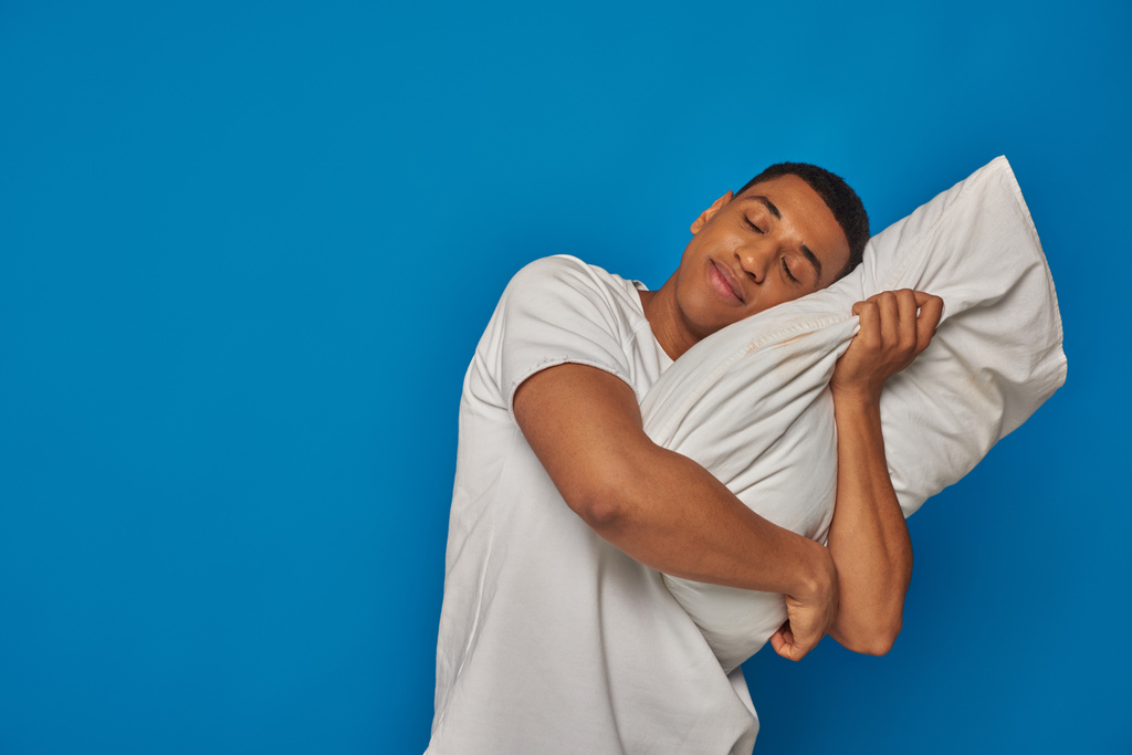 κουρασμένος αφροαμερικανός άντρας ξαπλωμένος στο μαξιλάρι και χαμογελαστός στο μπλε φόντο, νυσταγμένος άνθρωπος - Φωτογραφία, εικόνα