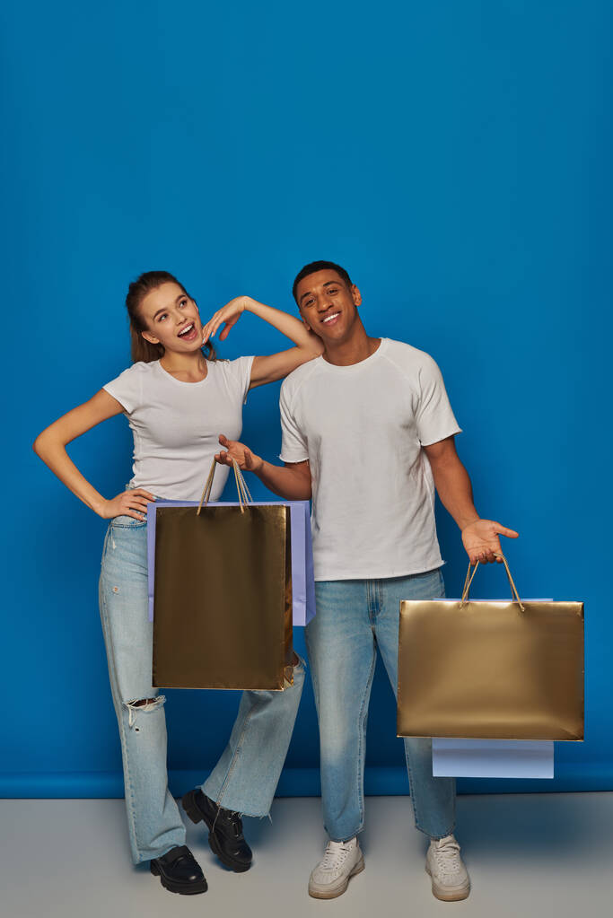 consumerism, happy interracial couple  holding shopping bags and standing together on blue backdrop - Photo, Image