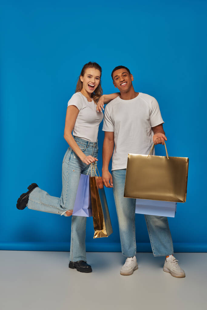 святкові продажі, щаслива міжрасова пара тримає сумки для покупок і стоїть на синьому фоні - Фото, зображення