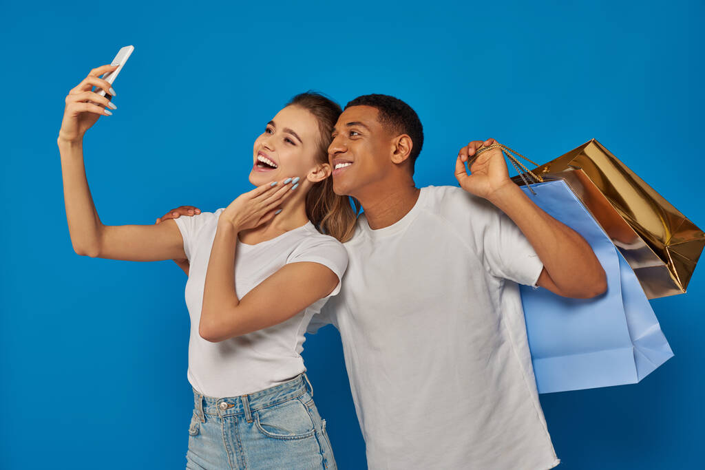 consumismo, feliz casal interracial segurando sacos de compras e tomando selfie no fundo azul - Foto, Imagem