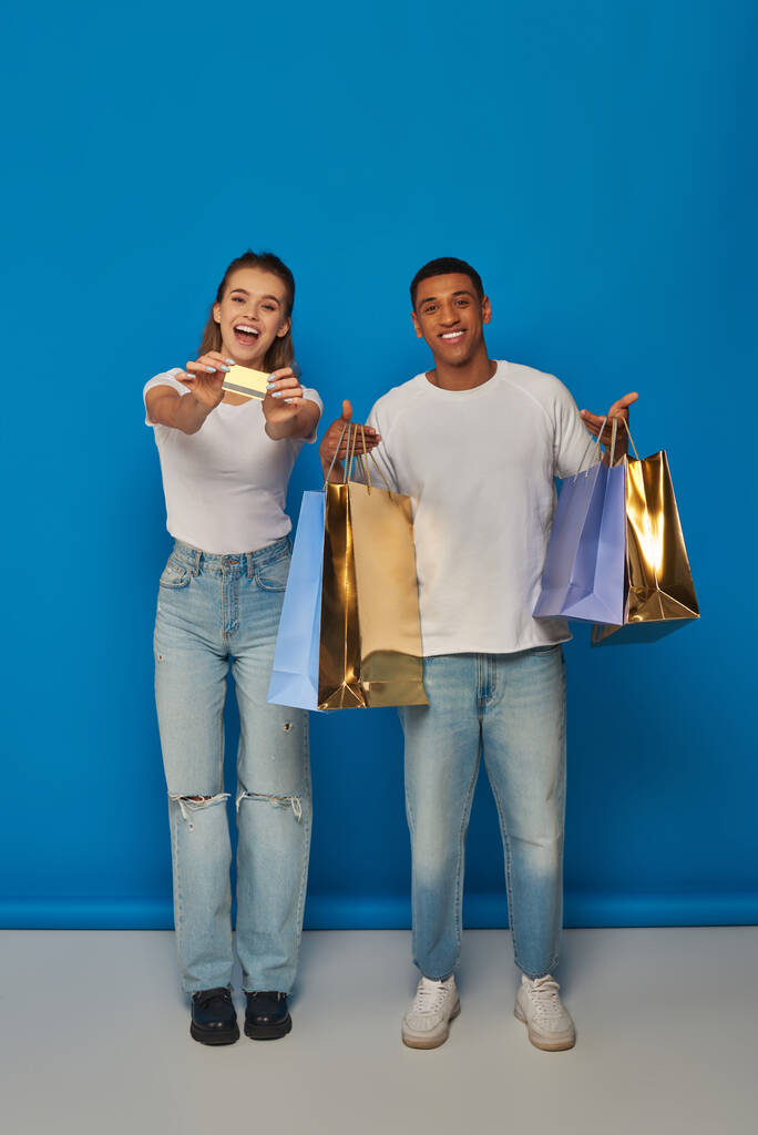 alegre pareja interracial sosteniendo bolsas de compras y tarjeta de crédito sobre fondo azul, consumismo - Foto, imagen