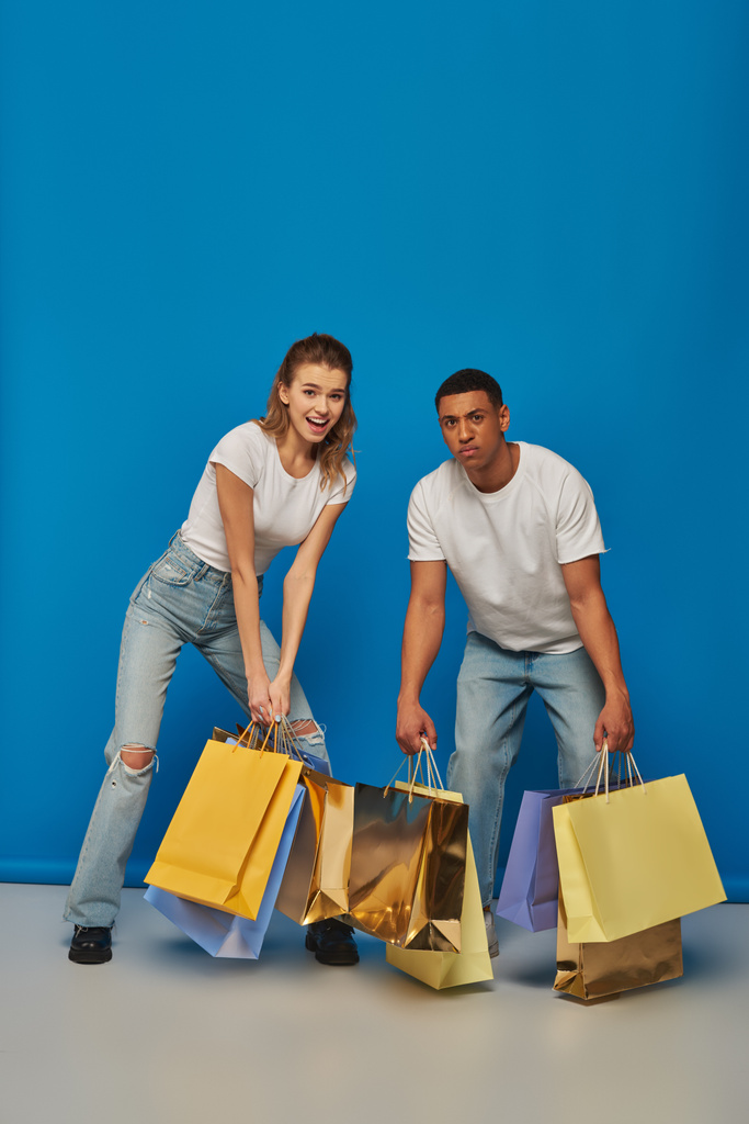 позитивный мультикультурный мужчина и женщина, держащие сумки для покупок на синем фоне, потребительства и продаж - Фото, изображение
