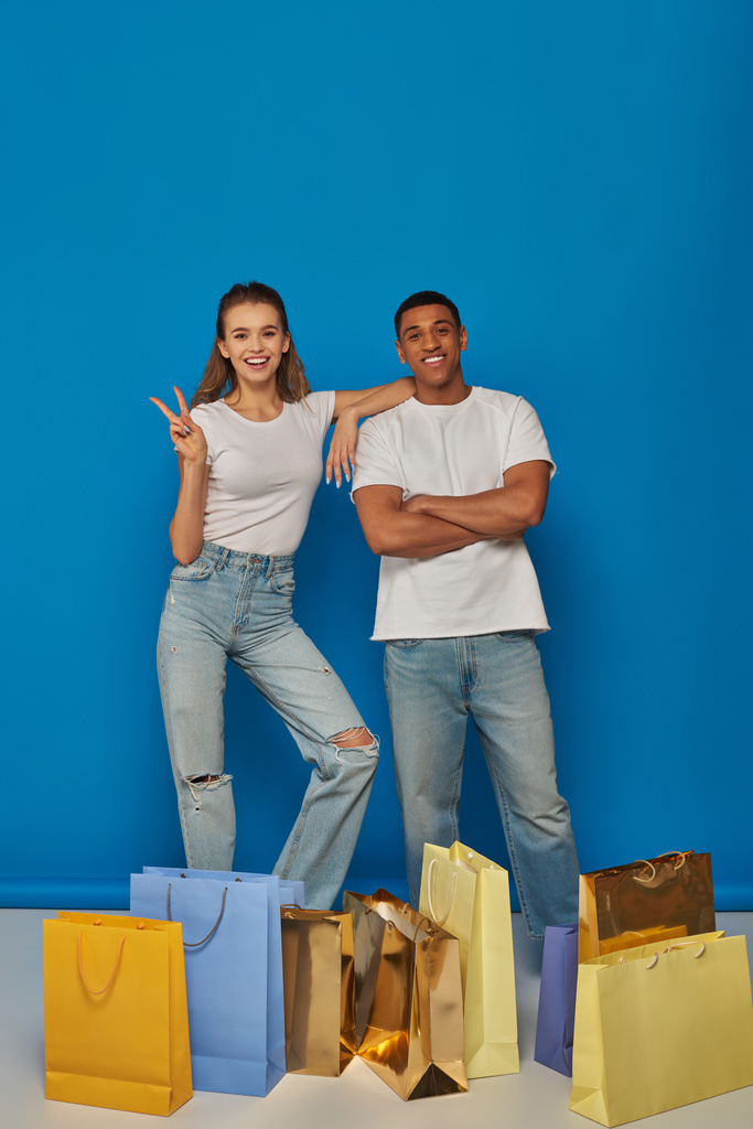 pozitivní multikulturní pár stojící v blízkosti nákupních tašek na modrém pozadí, ukazující mírové znamení - Fotografie, Obrázek