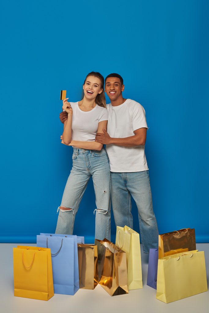 positiivinen monikulttuurinen pari seisoo ostoskassien lähellä ja pitää luottokorttia sinisellä taustalla - Valokuva, kuva