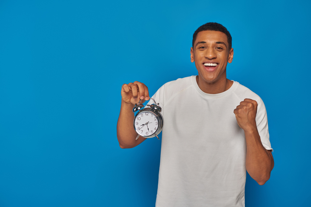 hombre afroamericano excitado con la boca abierta sosteniendo reloj despertador sobre fondo azul, expresión de la cara - Foto, imagen