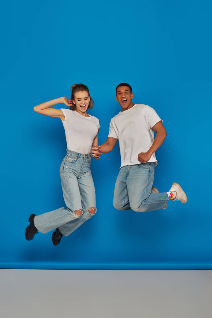 positive multicultural couple holding hands and jumping  on blue background, full length view - Photo, Image