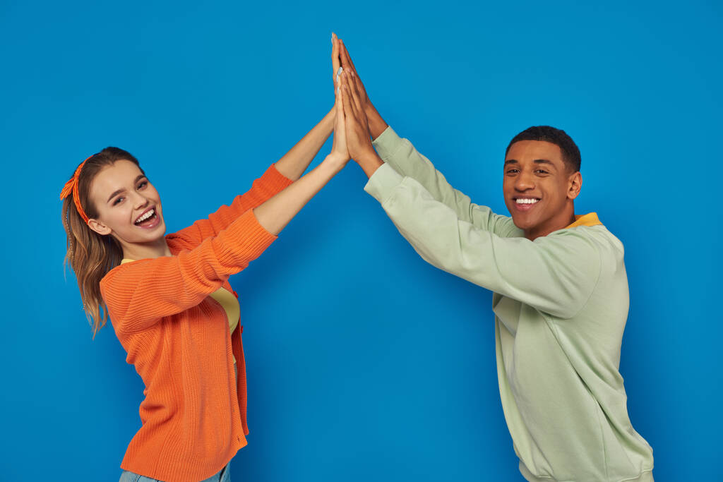 excitada pareja multicultural en atuendo casual dando alta cinco y sonriendo sobre fondo azul - Foto, Imagen