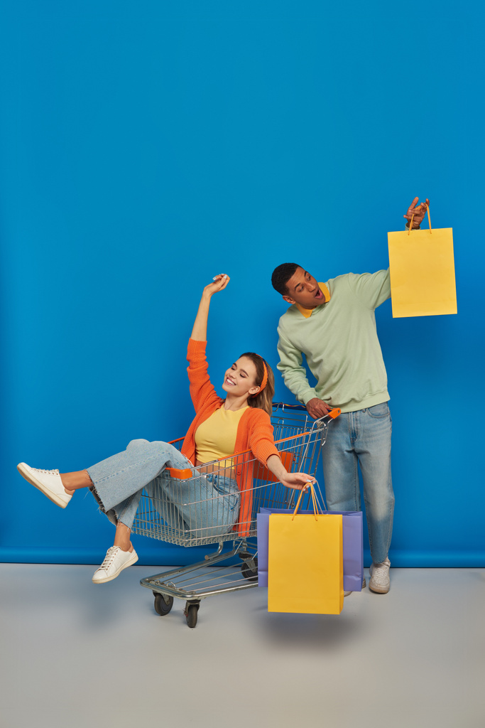 szczęśliwy Afroamerykanin mężczyzna i młoda kobieta w wózku sklepowym trzymając torby na niebieskim tle - Zdjęcie, obraz