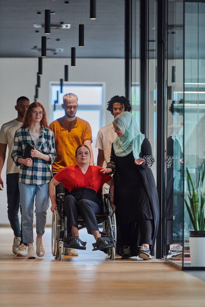 Monipuolinen ryhmä nuoria liikemiehiä, jotka kävelevät käytävällä modernin startupin lasisessa toimistossa, mukaan lukien pyörätuolissa oleva henkilö ja hijabia käyttävä nainen, jossa on dynaaminen yhdistelmä - Valokuva, kuva