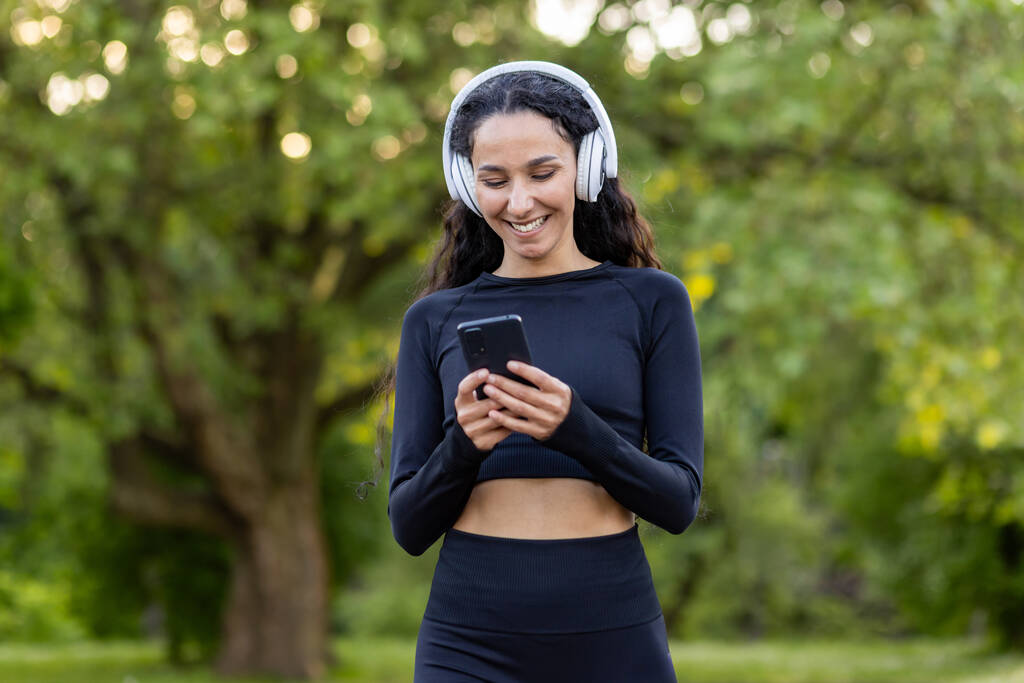 Úspěšná štíhlá latinskoamerická žena v parku během aktivního cvičení a jogging používá aplikaci smartphone, sportovkyně se usmívá ve sluchátkách, poslouchá audio podcasty a online rádio. - Fotografie, Obrázek