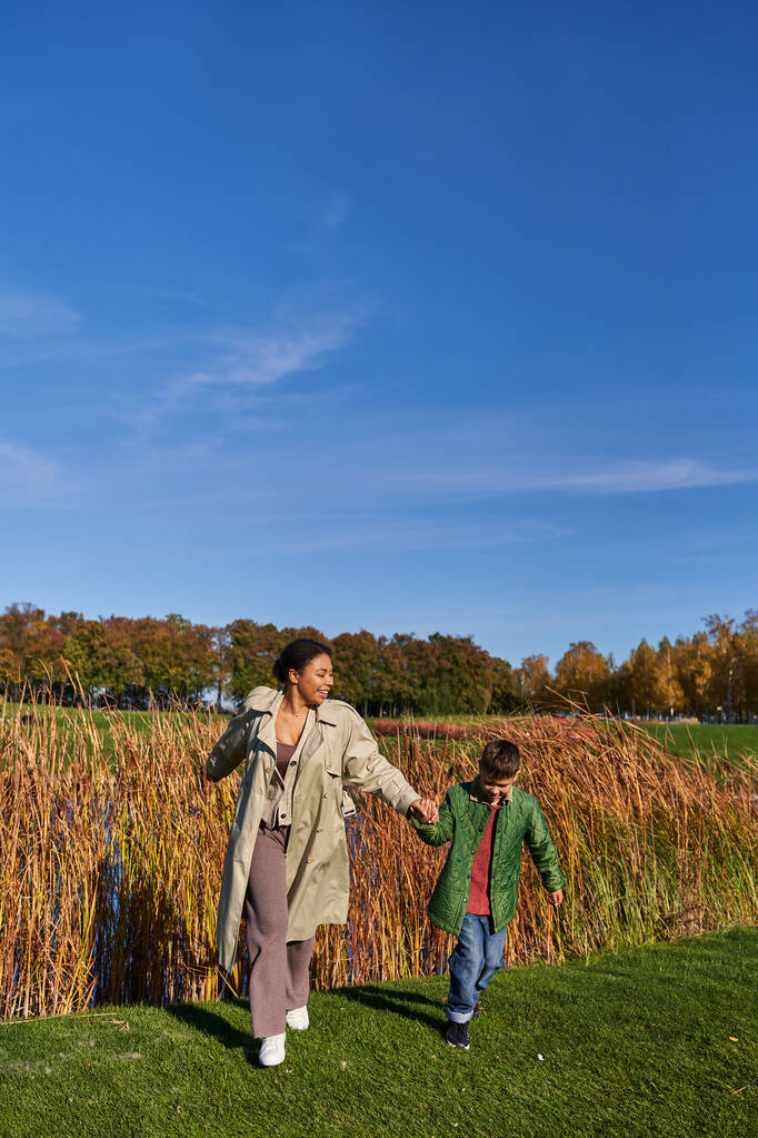 vinculación, personas y naturaleza, madre afroamericana feliz corriendo con su hijo, ropa de abrigo, otoño - Foto, imagen