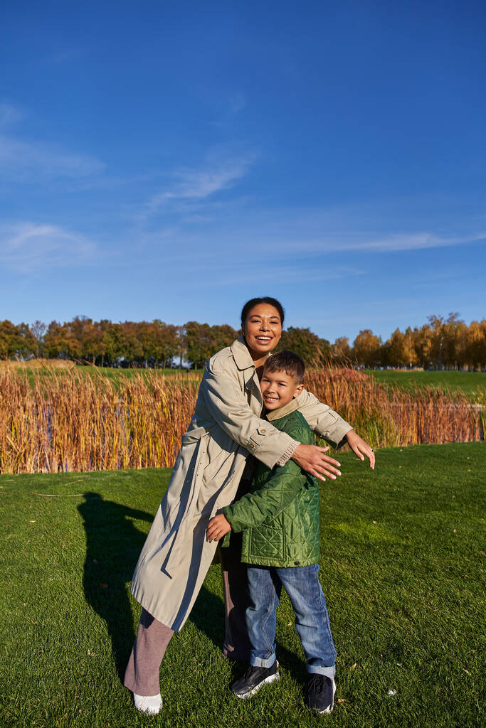 vinculación, naturaleza otoñal, madre afroamericana feliz abrazando al hijo, familia en ropa de abrigo, caída - Foto, Imagen