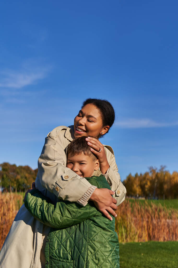 hechting, herfst natuur, gelukkig Afrikaans Amerikaans moeder omarmen zoon, familie in bovenkleding, vallen - Foto, afbeelding