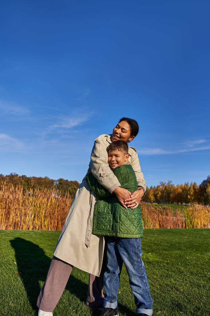 verbindende, herbstliche Natur, fröhliche afrikanisch-amerikanische Mutter umarmt Sohn, Familie in Oberbekleidung - Foto, Bild